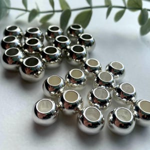 May include: A group of silver-toned metal beads with a hole in the center. The beads are round and smooth, and they are arranged in a loose pile.