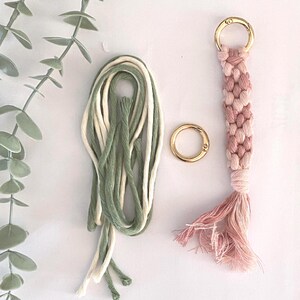 K&ouml;nnte beinhalten: Ein Makramee-Schl&uuml;sselanh&auml;nger mit einem geflochtenen Design in Rosa und Wei&szlig;, einem goldfarbenen Ring und einem B&uuml;ndel gr&uuml;ner und wei&szlig;er Makramee-Kordel. Der Schl&uuml;sselanh&auml;nger hat eine Quaste am unteren Ende. Ein zweiter goldfarbener Ring ist ebenfalls vorhanden.