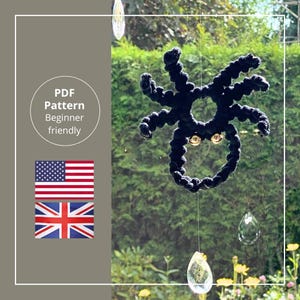 Op de afbeelding: Een zwarte, handgemaakte spinnendecoratie hangt voor een raam. De spin heeft goudkleurige ogen en is gemaakt van een dik, getextureerd materiaal. De tekst "PDF Pattern Beginner friendly" staat in een witte cirkel. Vlaggen van de VS en het VK zijn ook zichtbaar.