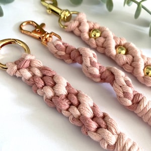 May include: Close-up of three macrame keychains in shades of pink and cream. Each keychain features a braided design with gold-colored hardware, including clasps and rings. Some keychains have gold beads.