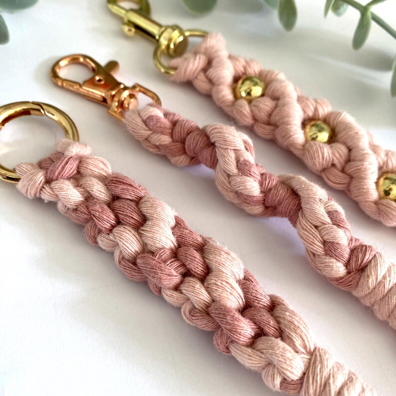 May include: Close-up of three macrame keychains in shades of pink and cream. Each keychain features a braided design with gold-colored hardware, including clasps and rings. Some keychains have gold beads.