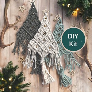 Puede incluir: Tres adornos de pared de macramé con forma de árbol de Navidad en gris oscuro, crema y verde salvia, cada uno con cuentas doradas y flecos. Un gráfico circular con el texto DIY Kit está superpuesto en el lado derecho de la imagen.