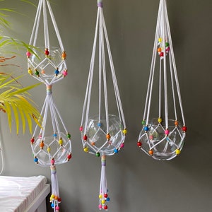 May include: Three white macrame plant hangers with glass globes and colorful wooden beads. The hangers are suspended from wooden rings and have tassels at the bottom.
