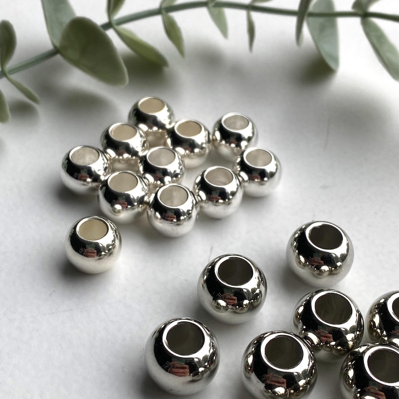 May include: A group of silver-toned metal beads with a hole in the centre. The beads are arranged in a circular pattern on a white surface.