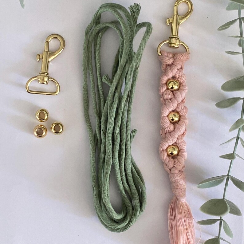 K&ouml;nnte beinhalten: Ein Makramee-Schl&uuml;sselanh&auml;nger-Set mit einem rosa gewebten Design, goldenen Perlen und einem goldenen Verschluss. Das Set enth&auml;lt auch gr&uuml;ne Kordel und goldfarbene Metallperlen. Der Schl&uuml;sselanh&auml;nger hat eine Quaste am Ende.