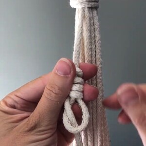 May include: A close-up of a hand tying a knot in a white macrame cord. The cord is attached to a wooden ring.