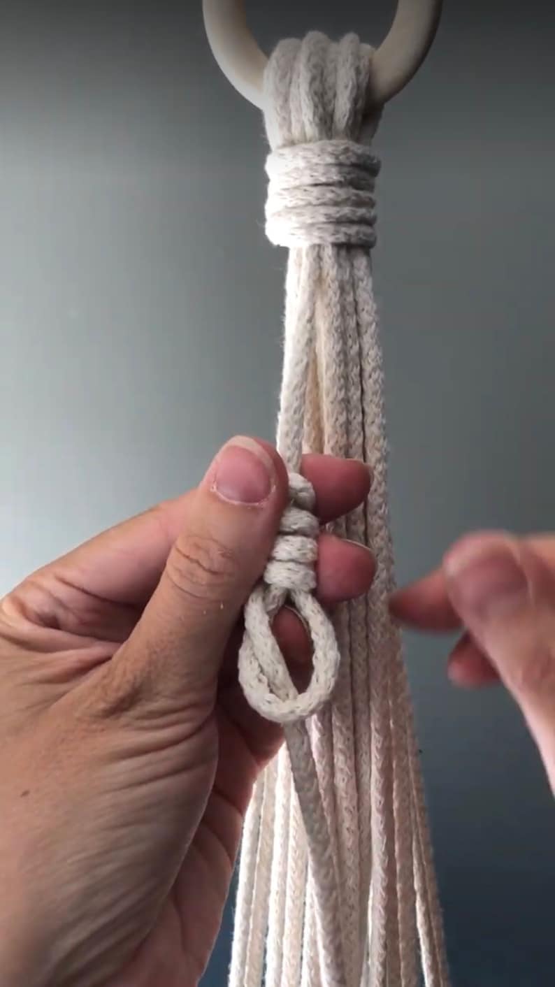 May include: A close-up of a hand tying a knot in a white macrame cord. The cord is attached to a wooden ring.