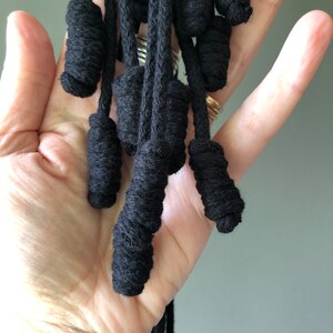 May include: A close-up of a black macrame tassel with multiple strands of cord tied into knots. The tassel is held in a person's hand.