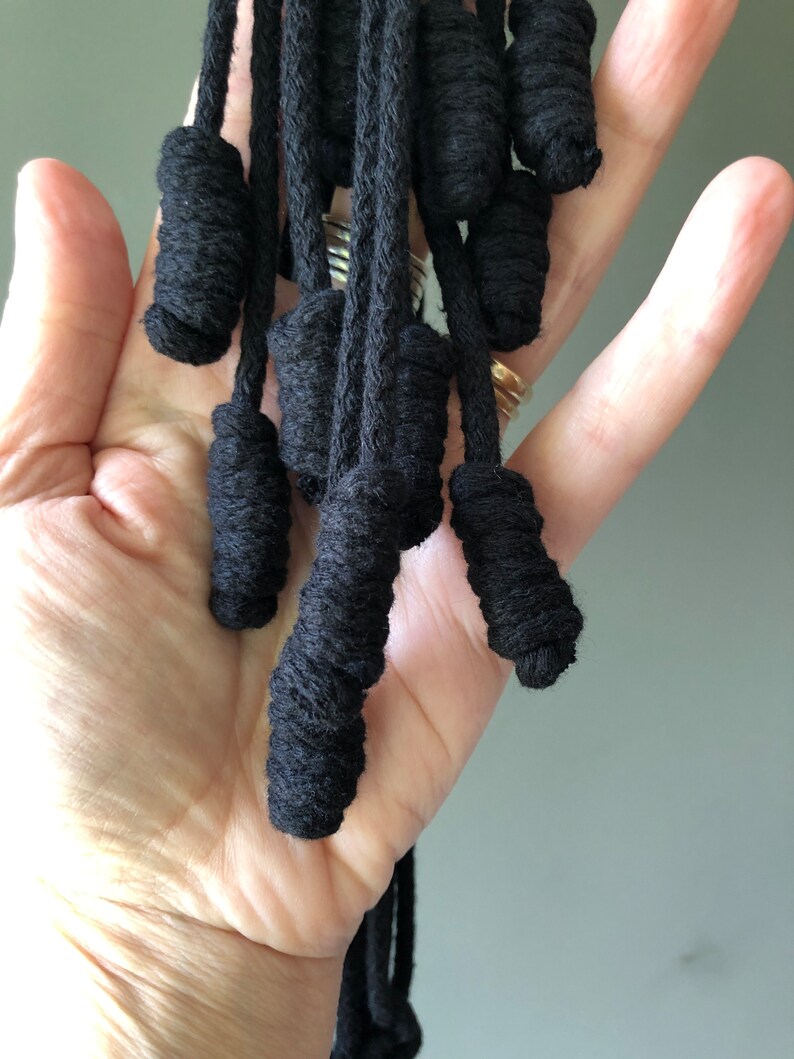 May include: A close-up of a black macrame tassel with multiple strands of cord tied into knots. The tassel is held in a person's hand.