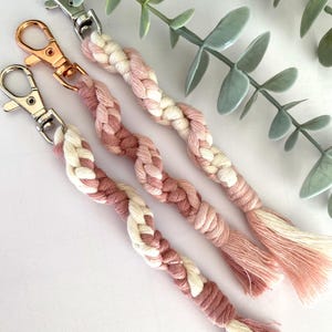 May include: Four macrame keychains in shades of pink and white, each with a metal clasp. The braided design transitions from light to darker pink, with a tassel at the end. The keychains are arranged diagonally against a white background.