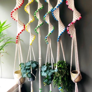 May include: Six macrame plant hangers with colourful beads hanging from a wooden dowel rod. The hangers are made of natural cotton rope and have a spiral design. Each hanger has a different colour of beads, including red, yellow, green, blue, and pink.