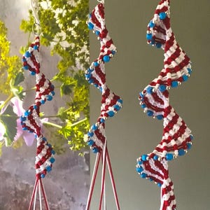 Op de afbeelding: Drie rode, witte en blauwe spiraalvormige macramé-oorbellen met blauwe kralen. De oorbellen hangen aan een witte achtergrond.