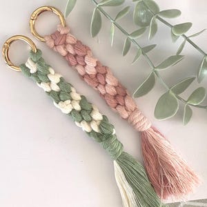 K&ouml;nnte beinhalten: Zwei Makramee-Schl&uuml;sselanh&auml;nger mit goldfarbenen Verschl&uuml;ssen. Einer ist gr&uuml;n und wei&szlig;, der andere rosa. Beide haben geflochtene Designs und Quastenenden. Die Schl&uuml;sselanh&auml;nger werden auf einer wei&szlig;en Oberfl&auml;che mit gr&uuml;nem Laub gezeigt.