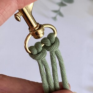 K&ouml;nnte beinhalten: Nahaufnahme eines Makramee-Projekts mit olivgr&uuml;ner Kordel, die an einem goldfarbenen Verschluss geknotet ist. Die Kordel ist eng gewebt und zeigt die Textur und die Details des handgefertigten Handwerks. Der Verschluss ist goldfarben.