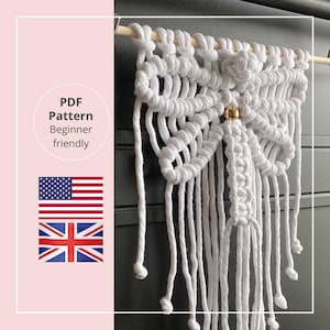 Op de afbeelding: Een witte macramé wandhanger met een gedetailleerd geweven ontwerp, met een centrale gouden kraal en hangende franjes. De tekst "PDF Pattern Beginner friendly" is zichtbaar, samen met de Amerikaanse en Britse vlaggen. De macramé is gemonteerd op een houten deuvel.