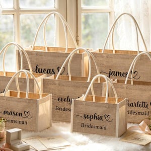 May include: A collection of beige burlap tote bags with white handles, personalized with names and titles like "Bridesmaid" and "Groomsman" in black script. The bags are arranged near a window, with flowers and a ring box visible.