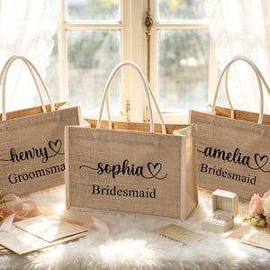 May include: Three burlap tote bags with white handles. Each bag has a name and title in black script: "henry Groomsma", "sophia Bridesmaid", and "amelia Bridesmaid". A ring box and flowers are in front of the bags.