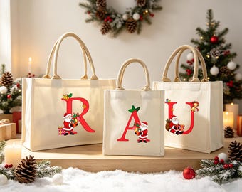 May include: Three off-white canvas tote bags with beige rope handles, each featuring a large red letter R, A, or U. Each letter is decorated with a graphic of Santa Claus holding gifts and holly leaves, creating a festive holiday design.