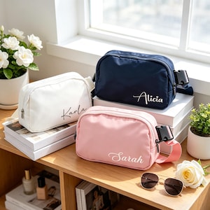 May include: Three personalized fanny packs in white, navy blue, and pink. Each pack has a name in cursive script: "Karla", "Alicia", and "Sarah". The bags are on a wooden surface with books, flowers, and sunglasses.