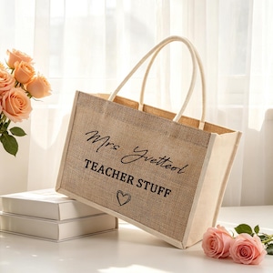 Pu&ograve; includere: Una borsa tote rettangolare in juta con due manici, caratterizzata dal testo stampato in nero Mrs Yvetteol, TEACHER STUFF e una piccola icona a forma di cuore. La borsa ha una trama intrecciata color marrone naturale e pannelli laterali bianchi.