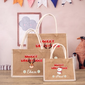 May include: Three burlap tote bags with white handles and red "Merry Christmas" text. The largest bag has a reindeer design and the name "Chase" printed below. The middle bag has a reindeer design. The smallest bag has a snowman design and the name "Paul" printed below.