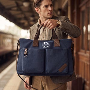 May include: A navy blue canvas messenger bag featuring two large front pockets, brown leather-style accents, and a metal D-ring. The bag is personalised with a white monogram logo containing the letter D and the name DANIEL in a rectangular frame.
