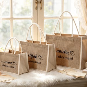 May include: Three burlap tote bags with white handles and trim are displayed. Each bag has a name and title printed in black script. The bags are on a white surface with flowers and a window in the background. The bags are for a wedding.