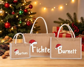 May include: Three rectangular tan jute tote bags with white handles. Each bag features a white name printed in a script font, topped with a small red and white Santa hat graphic. The names are Martens, Fichte, and Burnett, displayed in varying sizes.
