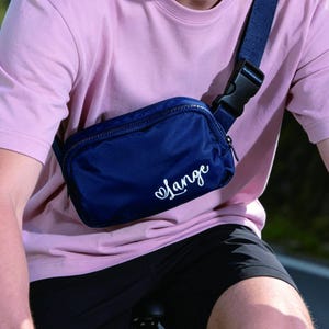May include: A navy blue crossbody bag with the word "Lange" in white script. The bag has a zippered closure and an adjustable strap with a black buckle. The person is wearing a pink t-shirt and black shorts.
