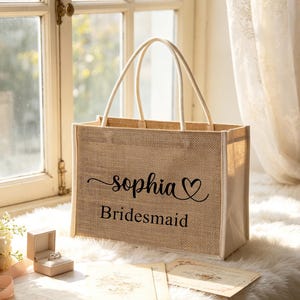 May include: A beige burlap tote bag with cream-colored handles. The bag features the name "Sophia" in cursive with a heart symbol, and the word "Bridesmaid" in a simple font. A ring box and a bouquet of roses are also visible.