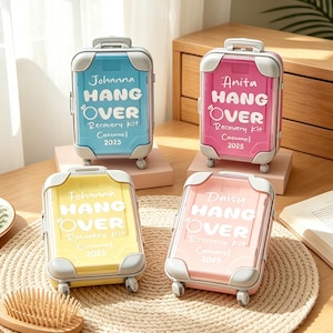 May include: Four miniature, colorful suitcase-shaped boxes. Each has a different name printed on it: Johanna, Anita, Fohanna, and Daisy. The text "HANG OVER Recovery Kit" is printed on each box, along with "Carmel 2025". The suitcases are blue, pink, yellow, and light pink.