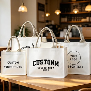 Peut inclure: Quatre sacs fourre-tout en toile blanche avec des poignées beiges sont présentés sur une table en bois. Chaque sac présente du texte personnalisé ou un logo en noir. Un sac dit "CUSTOM YOUR PHOTO", un autre dit "CUSTOM SECOND TEXT HERE".
