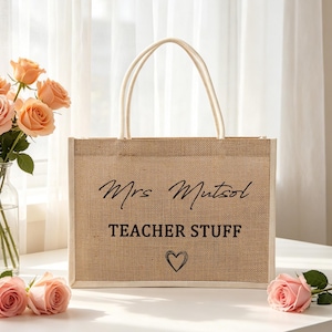Pu&ograve; includere: Una borsa tote rettangolare in juta color marrone chiaro con due manici bianchi. La parte anteriore presenta il testo nero Mrs Mutsol, le parole TEACHER STUFF in un carattere sans-serif in grassetto e un piccolo cuore disegnato a mano.