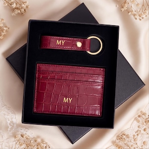 May include: A dark red crocodile-embossed card holder and a matching keychain with a gold-tone ring, both featuring the gold-stamped letters MY. The items are presented in a black gift box with a fitted insert, showcasing a textured finish.