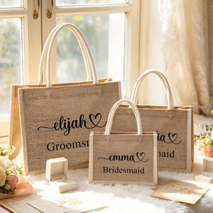 May include: Three beige burlap tote bags with white handles, personalized with names and titles in black script. The largest bag reads "elijah" and "Groomsmen". The medium bag says "tia" and "Maid". The smallest bag says "emma" and "Bridesmaid".