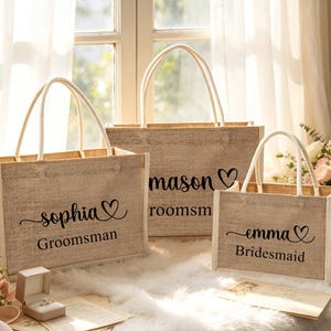May include: Three burlap tote bags with white handles and trim. The bags are personalized with names and titles in black script: "Sophia Groomsman", "Mason Groomsman", and "Emma Bridesmaid". A ring box and flowers are in the foreground.
