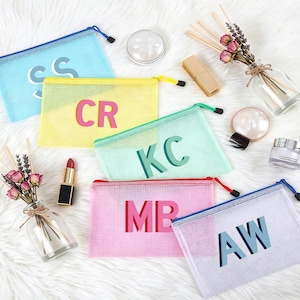 Puede incluir: Una colección de coloridas bolsas con cremallera de malla, cada una con una inicial diferente. Las bolsas son de color azul, amarillo, verde, rosa y blanco. También se ve maquillaje y un pequeño jarrón con flores.