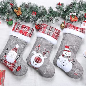 May include: Three grey Christmas stockings with names, Santa, reindeer, and snowman designs. Each stocking has a red and white plaid cuff with white faux fur trim. Festive holiday decorations.