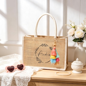 Puede incluir: Un bolso rectangular de yute color canela con dos asas blancas y un dise&ntilde;o de corona impreso en negro con el nombre Amelia en el centro. Una borla multicolor en tonos rosa, amarillo y azul cuelga de una de las asas del bolso.