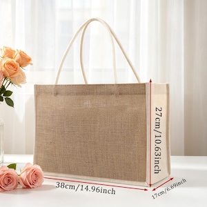 Pu&ograve; includere: Una borsa tote rettangolare in juta con pannelli laterali in tessuto bianco e due manici superiori bianchi. La borsa misura 38 cm di larghezza, 27 cm di altezza e 17 cm di profondit&agrave;, come indicato dalle frecce di misurazione rosse.