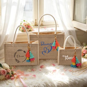 Puede incluir: Tres bolsas de mano de yute beige con asas blancas, cada una adornada con una borla colorida y texto personalizado. La bolsa más grande tiene "Dumas" en azul, la del medio "Olivia" en una corona y la más pequeña "Titan".