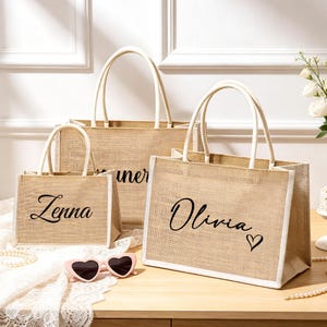 May include: Three rectangular jute tote bags with white trim and top handles. The bags feature black script text reading Zenna and Olivia, with a small heart icon next to the name Olivia. A pair of pink heart-shaped sunglasses rests in front.