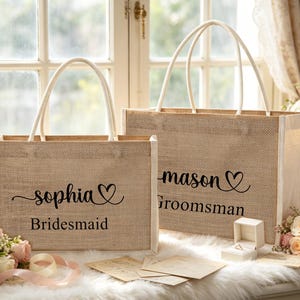 May include: Two beige burlap tote bags with white handles. One bag reads "Sophia" with a heart and "Bridesmaid" underneath. The other bag reads "mason" with a heart and "Groomsman".