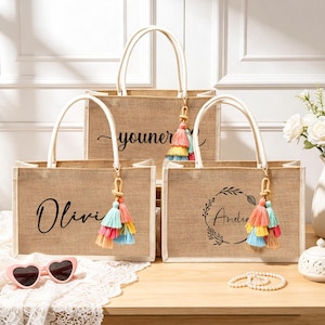 Puede incluir: Tres bolsos rectangulares de yute con asas blancas y borlas coloridas. Los bolsos presentan nombres y dise&ntilde;os impresos en negro, incluyendo los nombres Olivia y Amelia, y un gr&aacute;fico de corona floral. Tienen una textura de yute natural.