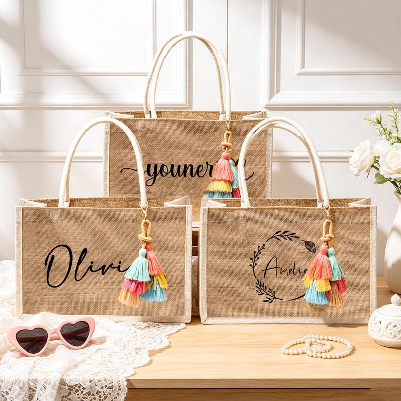 Puede incluir: Tres bolsos rectangulares de yute con asas blancas y borlas coloridas. Los bolsos presentan nombres y dise&ntilde;os impresos en negro, incluyendo los nombres Olivia y Amelia, y un gr&aacute;fico de corona floral. Tienen una textura de yute natural.