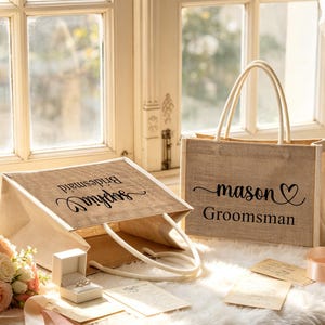 May include: Two burlap tote bags with white handles. One bag reads "Sophia Bridesmaid" and the other reads "mason Groomsman" with a heart. A ring box and flowers are in the foreground.