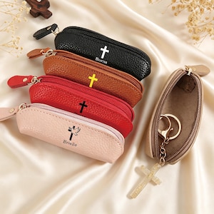 May include: A collection of five small, zippered, pebble-textured pouches in black, brown, red, and light pink. Each pouch features a cross graphic, with some including the names Burns or Brielle, and one open beige pouch containing a gold keychain with a cross.