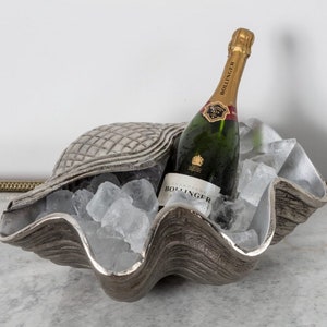 May include: A silver-colored clam shell ice bucket holding ice and a green champagne bottle with a gold foil top. The clam shell has a textured surface and is filled with ice cubes, keeping the bottle chilled.
