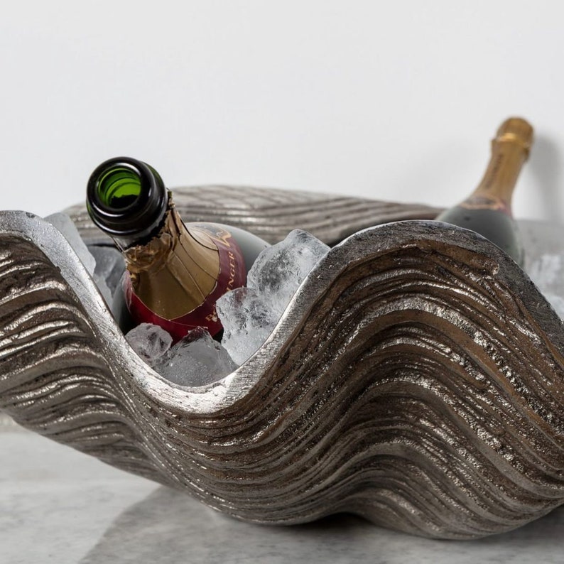 May include: A silver-colored, clam-shaped ice bucket holds two champagne bottles and ice cubes. The bucket has a textured, wavy design. One bottle is open, and the other is closed.