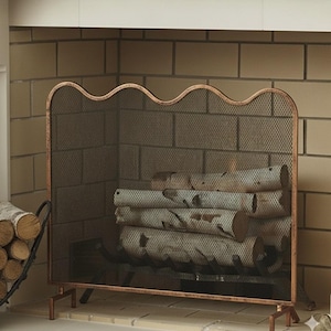 May include: A copper-colored fireplace screen with a wavy top edge and mesh screen. The screen is in front of a brick fireplace and contains several logs. A black metal log holder is visible on the left side of the image.